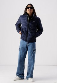 Jachetă puffer bleumarin cu închidere prin fermoar și model matlasat horizontal, purtată peste o cămașă neagră. Însoțită de blugi baggy de culoare albastru deschis și adidași albi.