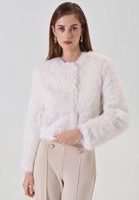 Weiße Kunstpelzjacke mit kurzem Schnitt, rundem Ausschnitt und langen Ärmeln; besitzt eine weiche, flauschige Textur. Kombiniert mit beigen, hoch taillierten Hosen.