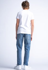 Camiseta blanca de manga corta combinada con jeans azul claro con detalles en los bolsillos, y llevada con zapatillas blancas. Vista trasera.