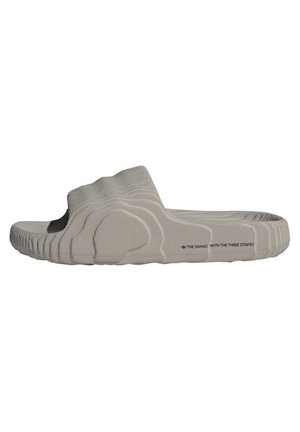 Lichtgrijze slidesandaal met een gestructureerde, golvende zool en bovenwerk, met de tekst "THE BRAND WITH THE THREE STRIPES" nabij de hiel.