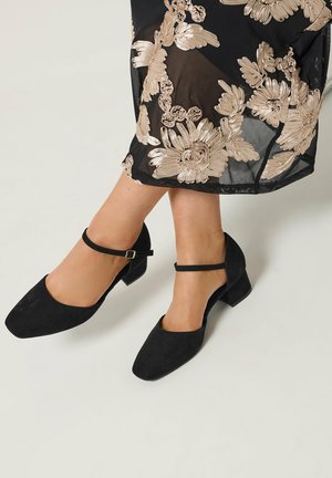 Beny i sorte ruskinds Mary Jane sko med lave hæle, kombineret med en gennemsigtig sort nederdel med store beige blomsterbroderier.
