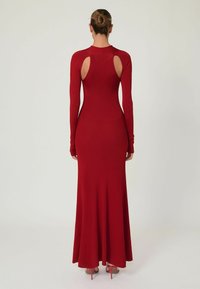 Frau mit einem Dutt, die ein enges, langes rotes Kleid mit ausgeschnittenen Schultern und High-Heels trägt, steht vor einem schlichten Hintergrund.