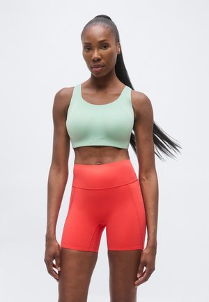 Atlétikus nő, hosszú lófarokkal, világoszöld sportmelltartót és élénk korall magas derekú biciklis rövidnadrágot visel, fehér háttér előtt.