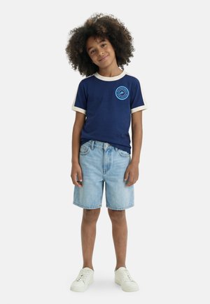 Child with curly hair wearing a navy blue t-shirt with logo, light denim shorts, and white sneakers, standing on a white background.