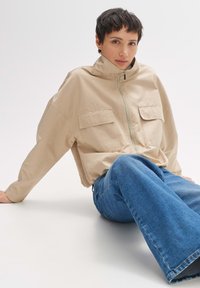 Beige Zip-Jacke mit hohem Kragen, zwei Fronttaschen und lockerem Schnitt; kombiniert mit ausgestellten blauen Jeans.