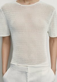 Vit stickad topp med korta ärmar och ett löst, öppet vävmönster, rund halsringning och rak fåll, tillsammans med ljusa byxor.