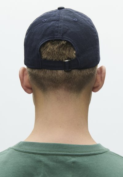 Kurzhaarige Person, die eine dunkelblaue Baseballkappe rückwärts trägt und ein grünes Rundhalsshirt anhat, von hinten vor einem schlichten Hintergrund betrachtet.