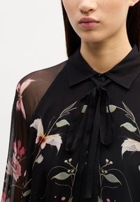 Blouse noire avec un motif floral composé de fleurs roses et blanches. Les manches transparentes et le détail du col noué rehaussent son apparence élégante.