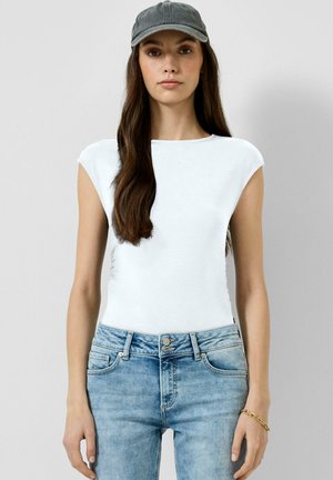 Femme portant un haut blanc sans manches, un jean bleu clair, une casquette grise et un bracelet en or, debout devant un fond gris uni.