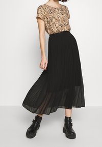 Beige top s černým květinovým vzorem, kombinovaný s černou plisovanou maxi sukní. Model má na sobě černé kotníkové boty se stahovacími pásky.