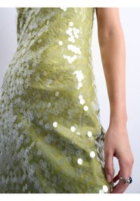 Gros plan d'une main avec une bague argentée tenant un tissu à sequins verts scintillants d'une robe sur un fond uni.