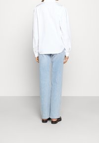 Chemise blanche à manches longues avec un col plat, associée à un jean bleu clair à jambes droites. Le tissu semble lisse et léger.