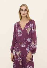 Ein pflaumenfarbiges Kleid mit langen, puffigen Ärmeln und einem V-Ausschnitt. Es ist mit Blumenmustern in Blau, Creme und Mauve verziert. Weicher, strukturierter Stoff.