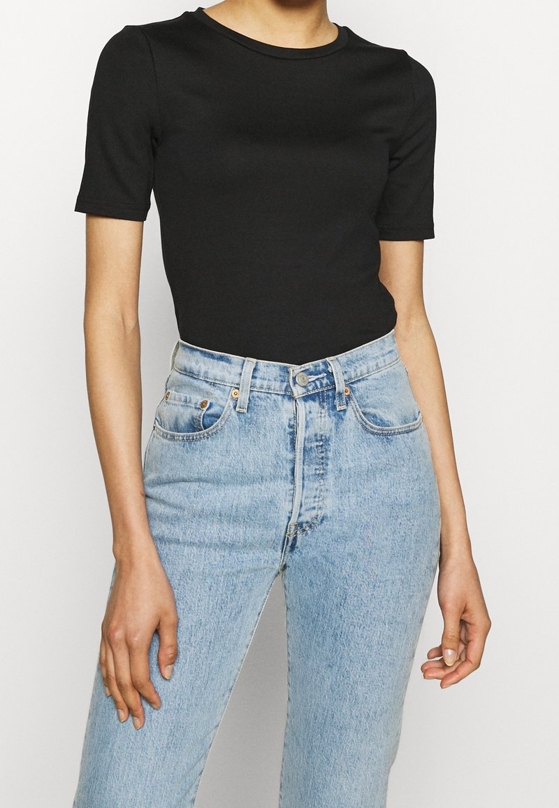 Buste féminin portant un t-shirt noir ajusté à manches courtes, rentré dans un jean en denim bleu clair taille haute avec une fermeture par bouton.