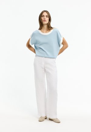 Frau steht mit den Händen hinter dem Rücken, trägt ein hellblaues kurzärmeliges Oberteil, weiße Hosen mit weitem Bein und beige geflochtene Ballerinas vor weißem Hintergrund.