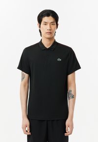 Svart polo t-shirt tillverkad av mjukt tyg, med krage, korta ärmar och en grön broderad krokodil-logga på vänster bröst.