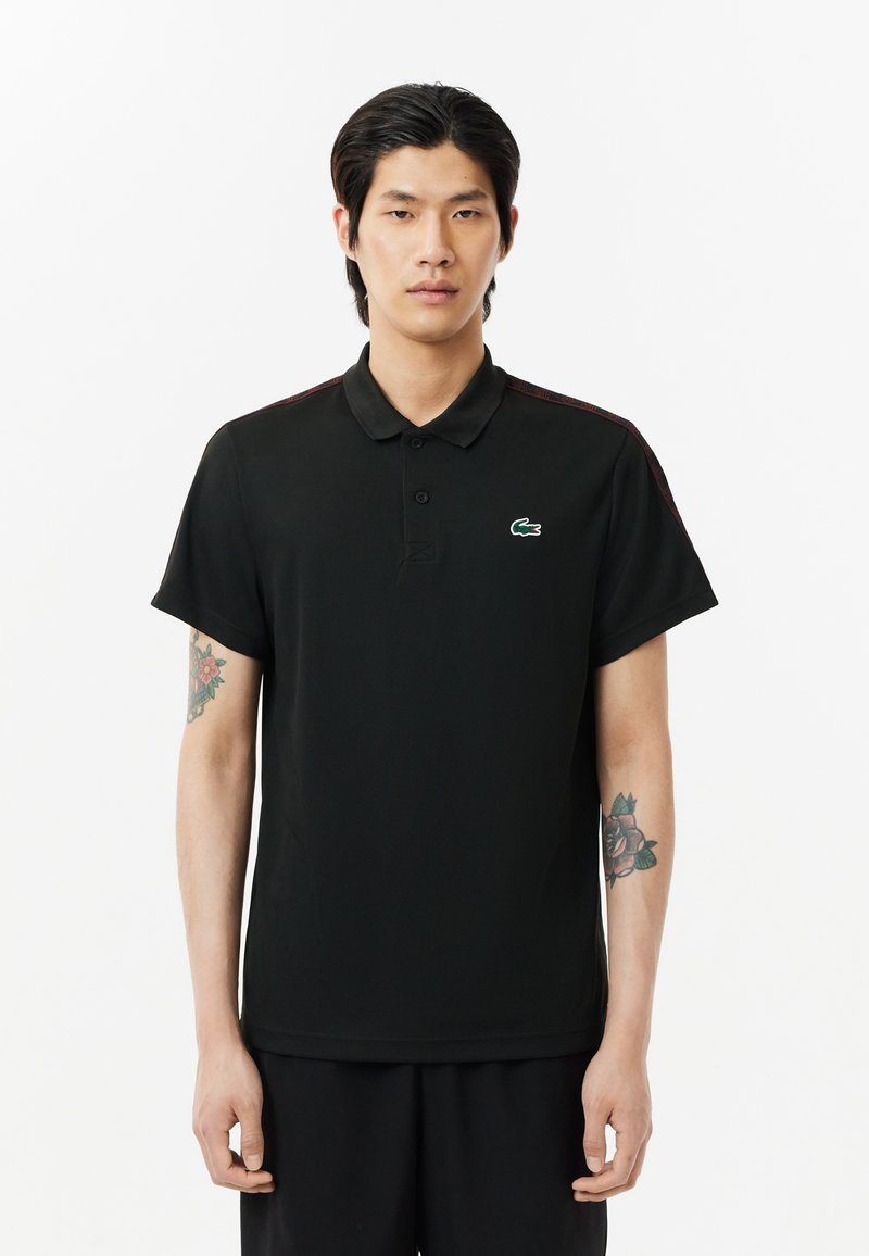 Schwarzes Polohemd aus glattem Stoff, mit Kragen, kurzen Ärmeln und einem grünen, gestickten Krokodil-Logo auf der linken Brust.