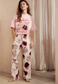 Lys rosa T-shirt med dessertgrafik og tekst; cremede bukser med farverige kager. Afslappet pasform og behagelig stil.
