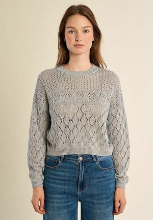 Giovane donna di fronte che indossa un maglione lavorato a maglia grigio chiaro con motivi e jeans blu su uno sfondo beige uniforme.