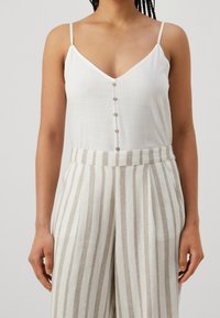 Top bianco senza maniche con scollatura a V e patta con cinque bottoni, abbinato a pantaloni ampi a vita alta a righe beige e bianche.