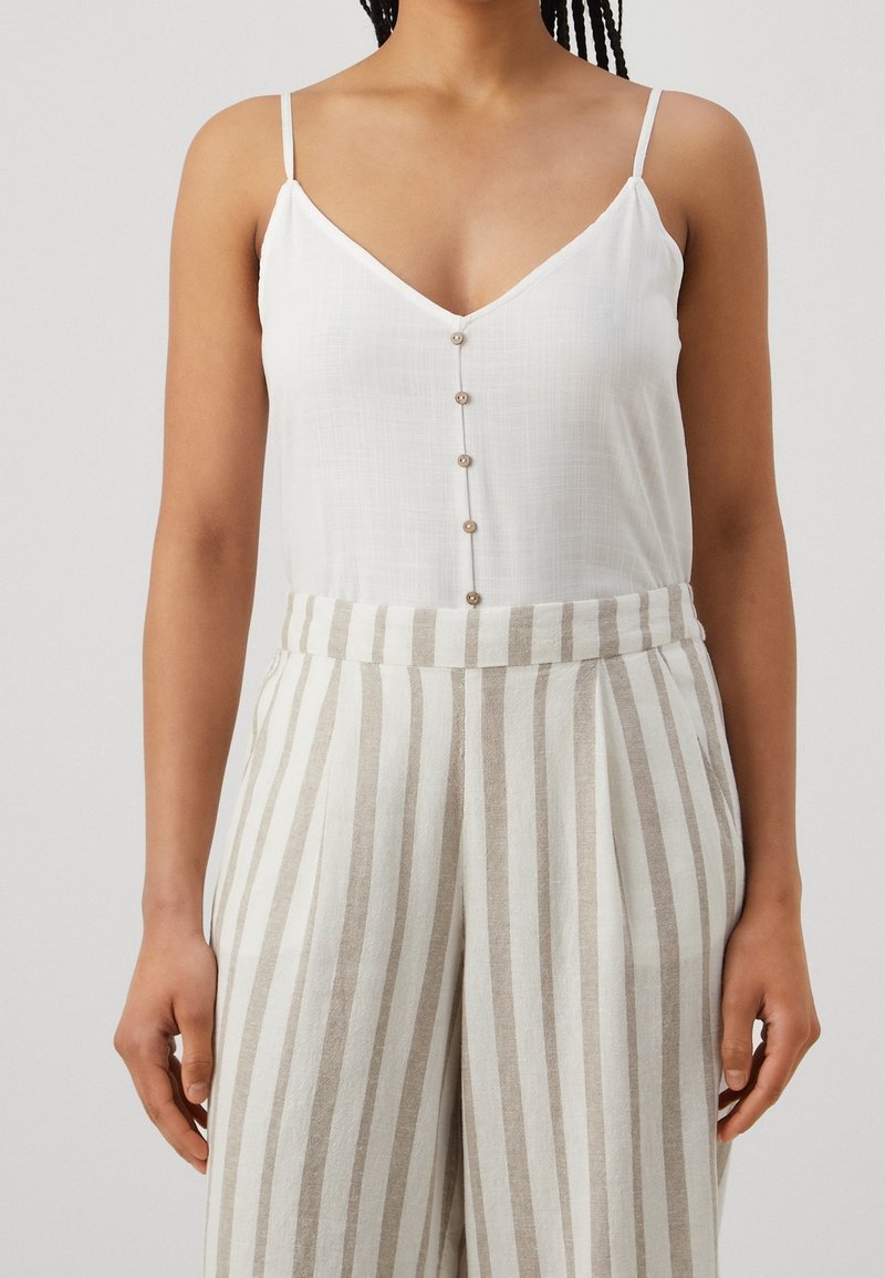 Top bianco senza maniche con scollatura a V e patta con cinque bottoni, abbinato a pantaloni ampi a vita alta a righe beige e bianche.