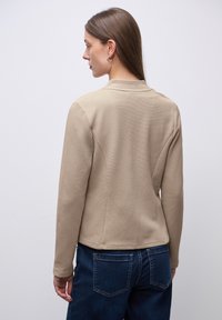 Veste beige texturée à manches longues et col, présentant un design ajusté et un dos sans coutures. Associée à un pantalon en denim bleu foncé.