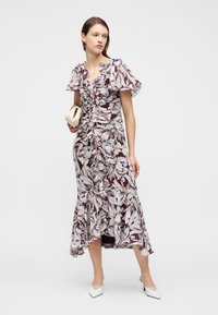 Νεαρή γυναίκα που φορά ένα μίντι λουλουδάτο φόρεμα με βολάν, κρατώντας μια μικρή μπεζ τσάντα χειρός και φορώντας λευκές μυτερές γόβες.