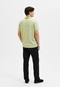 Camisa polo de manga corta para hombre en verde claro, confeccionada en tela suave, con un cuello tipo polo y un diseño de yugo en la parte posterior. Se lleva con pantalones negros.