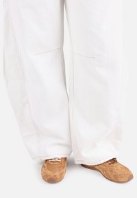 Pantalon blanc à jambes larges avec des détails de couture visibles, associé à des baskets en daim marron avec des lacets et des accents perforés.