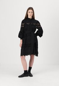Abito in pizzo nero con scollo alto, maniche lunghe a sbuffo e vita cintrata. Presenta intricati motivi a occhio di pernice e dettagli con orlo ondulato.