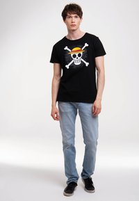 Black cotton t-shirt featuring a white skull and crossbones graphic with a yellow and red straw hat. Worn with light blue jeans and black shoes.