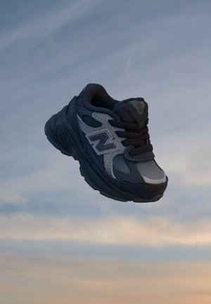 Baskets New Balance noires et grises avec lacets suspendues devant un ciel bleu et orange.