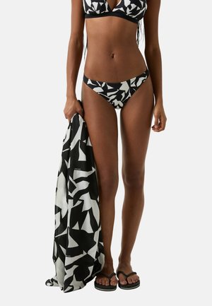 Bikini mit schwarz-weißer Musterung und dreieckigen Formen, kombiniert mit einem passenden Wickeltuch, das in einer Hand gehalten wird. Flache Sandalen sind an den Füßen getragen.