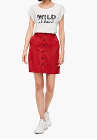 Mini gonna in suede rossa con chiusura frontale a bottoni e una texture liscia, abbinata a una T-shirt bianca con scritte nere. Sneakers bianche.