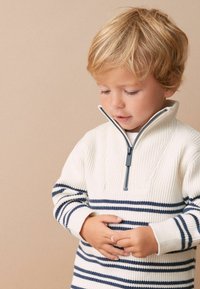 Suéter blanco de punto con rayas horizontales navy, cuello con cremallera alta, textura acanalada y mangas largas. Niño sosteniendo las manos juntas suavemente.