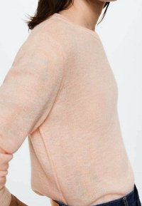 Hellrosa gestrickter Pullover mit Rundhalsausschnitt und langen Ärmeln. Weiche Textur, figurbetonte Form und minimale Nähte. Kombiniert mit dunklen Jeans.