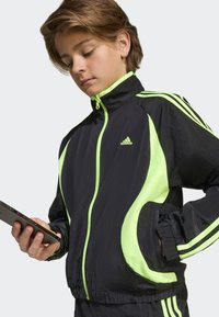 Crna sportska jakna s neonsko zelenim naglascima i prugama, s visokim ovratnikom, patentnim zatvaračem i teksturiranim tkanjem.