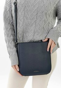 Frau trägt grauen Zopfmuster-Pullover und beige Hosen, hält eine schwarze Leder-Umhängetasche mit verstellbarem Riemen.