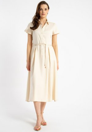 Kobieta w kremowej, krótkim rękawem, midi sukience na guziki z cienkim paskiem i złotych szpilkach z paskiem wokół kostki, stojąca na białym tle.