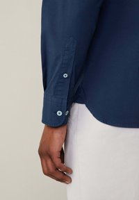 Camisa de manga larga azul marino con una textura suave, que presenta botones azul claro y un dobladillo curvado, parcialmente visible con una mano agarrando el puño.