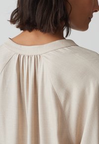 Beige blouse met een zachte textuur, geplooide details aan de achterkant en een eenvoudige kraag. De stof lijkt lichtgewicht en glad te zijn.