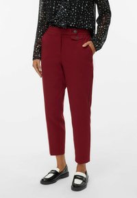 Mujer vistiendo pantalones burdeos de longitud hasta el tobillo con detalle de botones en la cintura, combinados con mocasines en blanco y negro y una blusa negra de gasa con estampado.