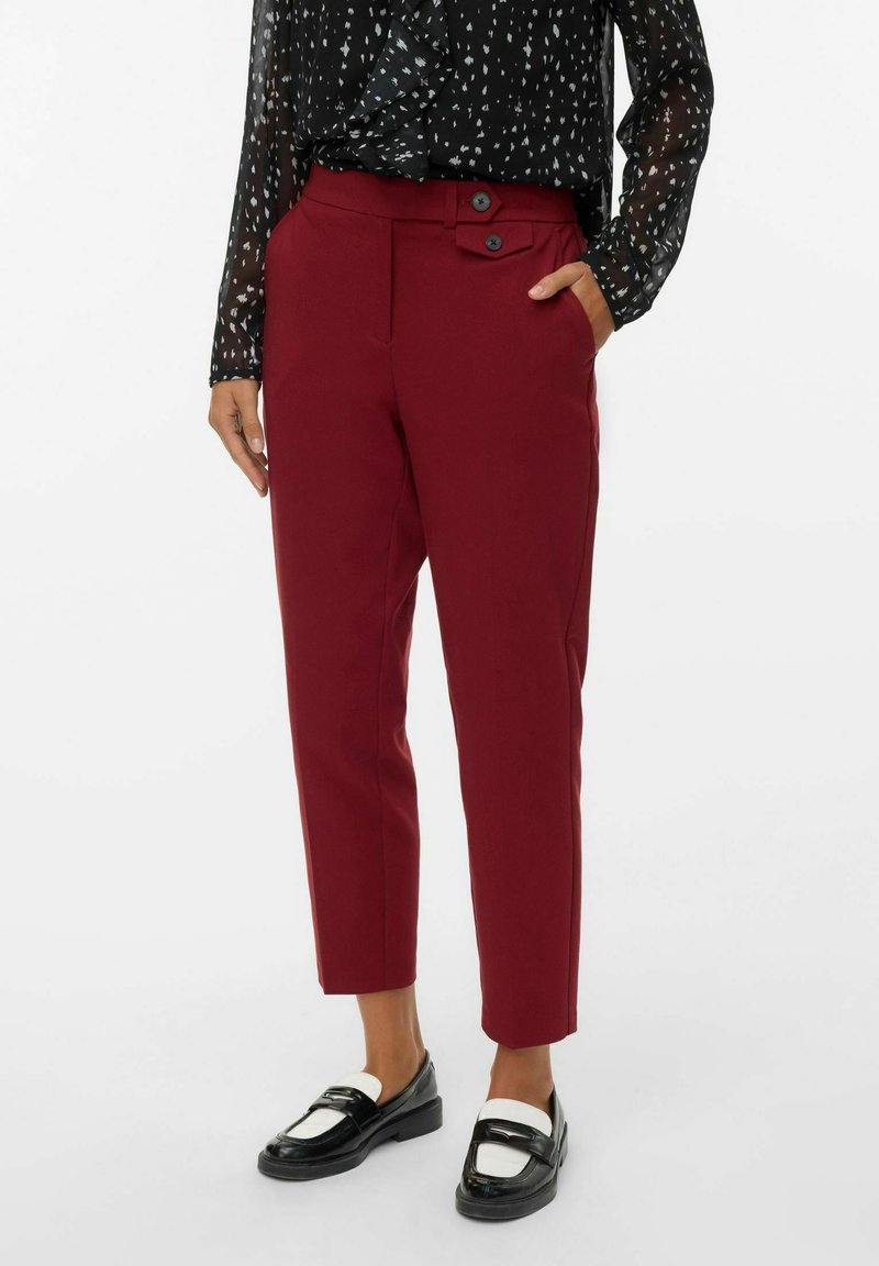 Mujer vistiendo pantalones burdeos de longitud hasta el tobillo con detalle de botones en la cintura, combinados con mocasines en blanco y negro y una blusa negra de gasa con estampado.