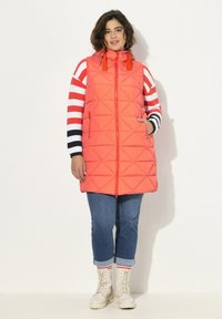 Gilet puffer trapuntato corallo con cappuccio, dotato di chiusura con zip e due tasche laterali. Il modello lo abbina a un maglione a righe e jeans.