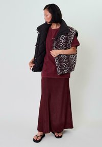 Bordeauxfarbenes Kleid mit lockerem Schnitt und langer Länge, kombiniert mit einer schwarzen, tiermuster-gesteppten Weste. Model trägt schwarze Flip-Flops.