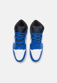 Ett par hög-top blåa, vita och svarta sneakers med blå snörningar och "Air Jordan"-loggor på den svarta tungan, sedd uppifrån på en vit bakgrund.