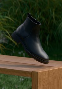 Schwarze Lederstiefelette mit glatter Oberfläche, elastischem Seitenpanel und rutschfester, strukturierter Sohle, platziert auf einem Holztisch.