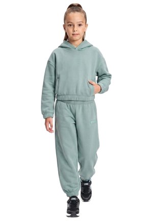Une jeune fille présente un sweat à capuche vert pâle et un pantalon de jogging assorti, avec des baskets noires, sur un fond blanc.