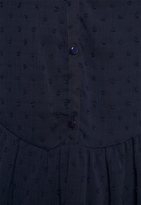 Vestido azul-marinho com tecido texturizado em trama, apresentando pequenos pontos elevados e fecho de botão. Inclui um design de saia franzida.