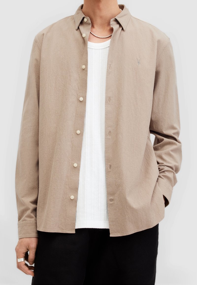 Camisa de botones marrón claro hecha de tejido texturizado, con un cuello clásico y botones beige, superpuesta sobre una camiseta blanca de canalé.